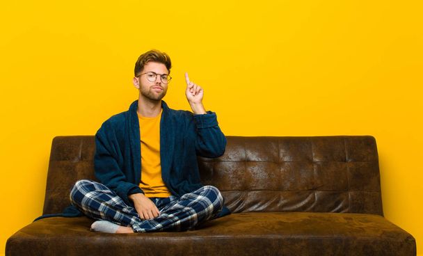 young man wearing pajamas feeling like a genius holding finger proudly up in the air after realizing a great idea, saying eureka . sitting on a sofa - Photo, Image