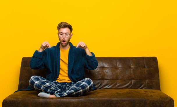 young man wearing pajamas feeling shocked, open-mouthed and amazed, looking and pointing downwards in disbelief and surprise . sitting on a sofa - Fotoğraf, Görsel