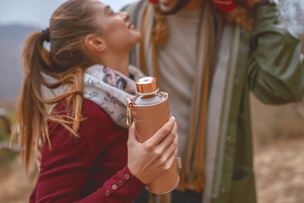Cute couple in love standing in nature and flirting. Man holding dog on shoulders while woman holding thermos with hot tea. Selective focus on thermos. Autumn season. - Photo, Image