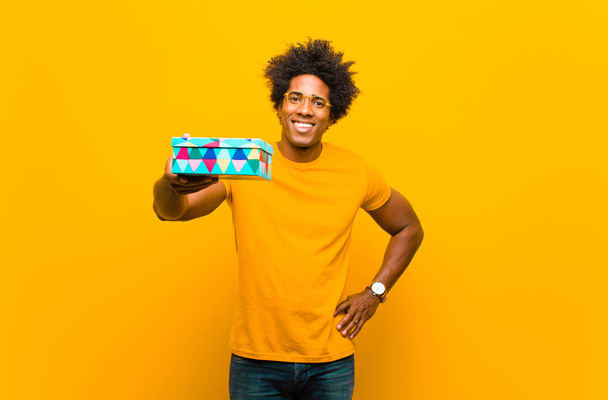 young african american man with a gift box against orange backgr - Foto, immagini