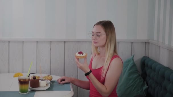 Όμορφο νεαρό κορίτσι κάθεται σε ένα τραπέζι σε ένα καφέ και τρώει γλυκά φρέσκα γλυκά από ένα πιάτο. - Πλάνα, βίντεο