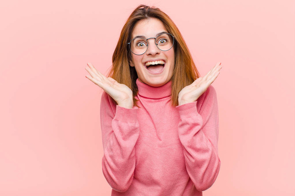 young pretty woman looking happy and excited, shocked with an unexpected surprise with both hands open next to face against pink background - Photo, image
