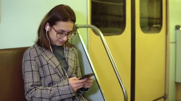Młoda kobieta słucha muzyki na słuchawkach z telefonem w rękach w pociągu metra. Dziewczyna jest w korespondencji telefonicznej. Stary wagon metra - Materiał filmowy, wideo