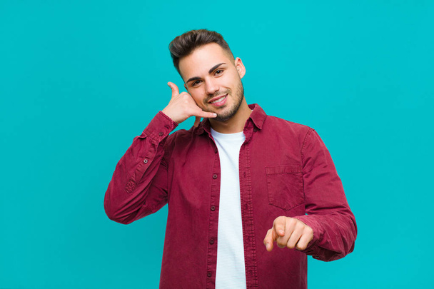 young hispanic man smiling cheerfully and pointing to camera while making a call you later gesture, talking on phone against blue background - Photo, image