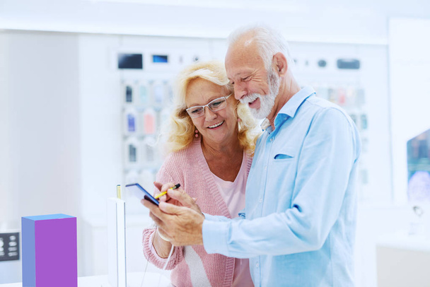 Linda sonriente pareja de ancianos de pie en la tienda de tecnología y probar el nuevo teléfono inteligente
. - Foto, Imagen