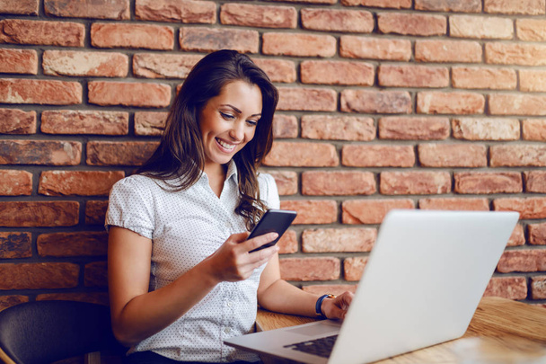 Занятая деловая женщина в рубашке сидит в кафе и пользуется смартфоном. На столе ноутбук. На заднем плане кирпичная стена
. - Фото, изображение
