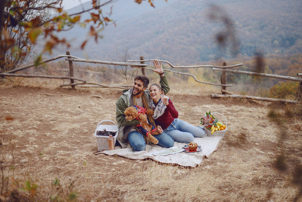 fröhliches attraktives multikulturelles Paar in lässiger Kleidung, das auf einer Decke in der Natur sitzt und winkt. Mann hält seinen Hund. Picknick im Herbst. - Foto, Bild