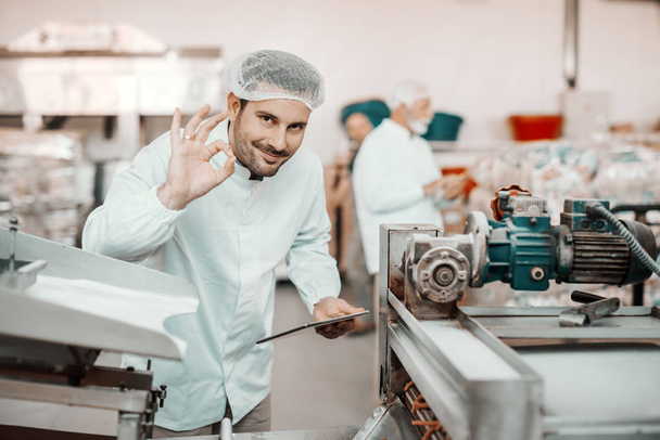 Young Caucasian smiling supervisor evaluating quality of food in food plant while holding tablet and showing okay sign. Man is dressed in white uniform and having hair net. - Photo, Image