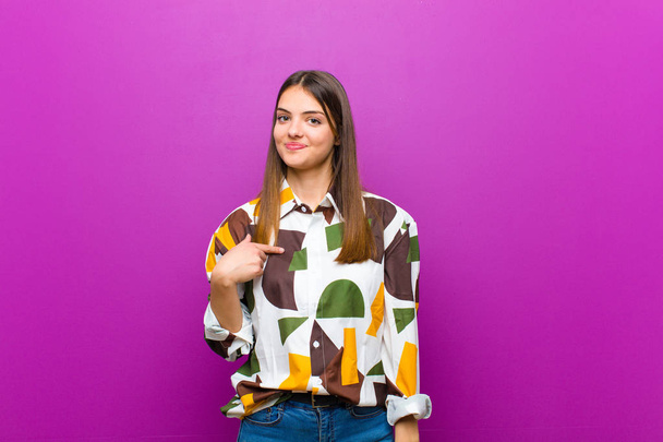 young pretty woman looking proud, confident and happy, smiling and pointing to self or making number one sign against purple background - 写真・画像