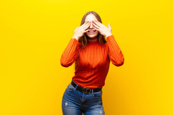 young pretty woman smiling and feeling happy, covering eyes with both hands and waiting for unbelievable surprise against orange background - Foto, Bild