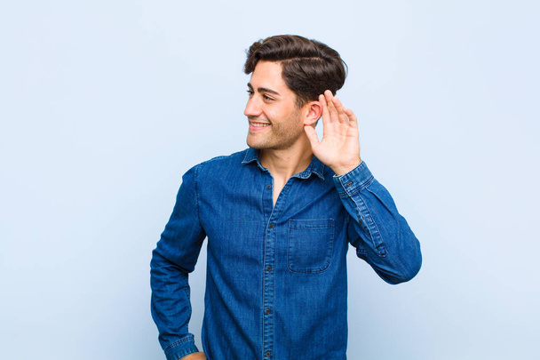 young handsome man smiling, looking curiously to the side, trying to listen to gossip or overhearing a secret against blue background - Photo, Image