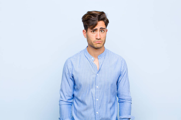 young handsome man feeling sad and whiney with an unhappy look, crying with a negative and frustrated attitude against blue background - Fotoğraf, Görsel