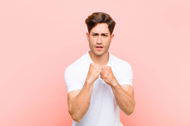 young handsome man looking confident, angry, strong and aggressive, with fists ready to fight in boxing position against pink background - Fotografie, Obrázek