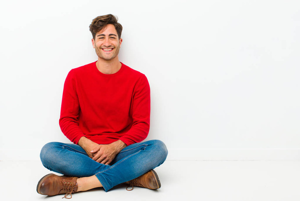 young handsome man smiling gleefully, feeling happy, satisfied and relaxed, with crossed arms and looking to the side sitting on the floor - Photo, image