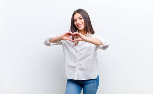 young pretty woman smiling and feeling happy, cute, romantic and in love, making heart shape with both hands isolated against white wall - Фото, изображение