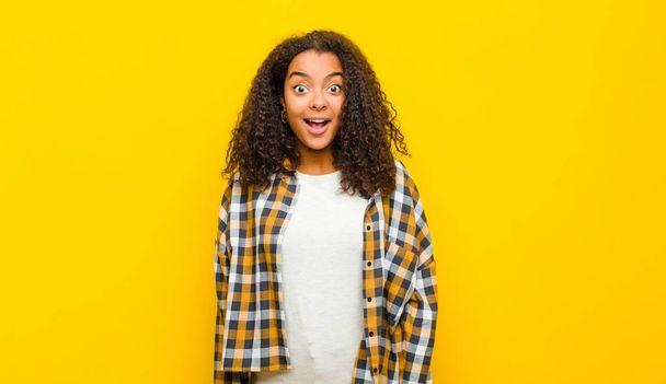 young pretty african american woman looking very shocked or surprised, staring with open mouth saying wow against yellow wall - Fotografie, Obrázek