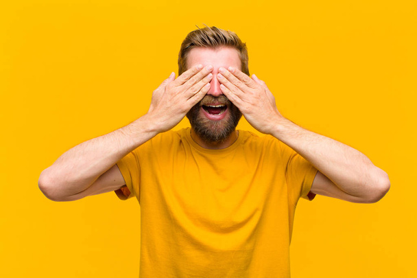 young blonde man smiling and feeling happy, covering eyes with both hands and waiting for unbelievable surprise against orange wall - Foto, Imagen