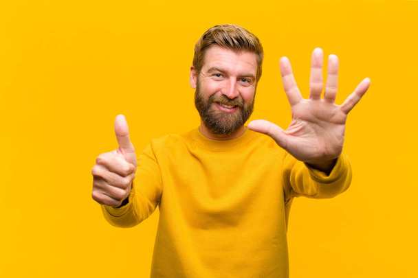 young blonde man smiling and looking friendly, showing number six or sixth with hand forward, counting down against orange wall - Fotoğraf, Görsel