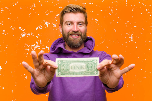 young blonde man with dollar bills or banknotes wearing a purple hoodie against damaged orange wall - Φωτογραφία, εικόνα