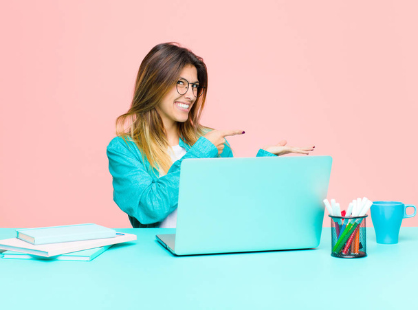 young pretty woman working with a laptop smiling cheerfully and pointing to copy space on palm on the side, showing or advertising an object - Photo, Image