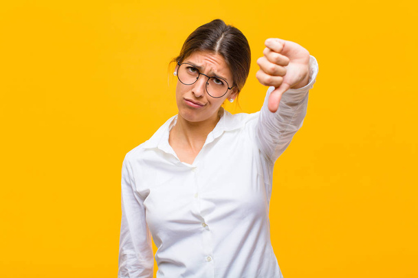young businesswoman feeling cross, angry, annoyed, disappointed or displeased, showing thumbs down with a serious look against orange wall - Foto, imagen