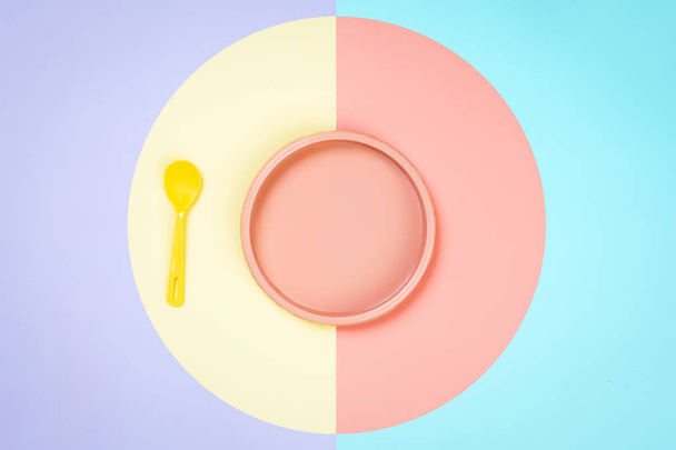 Minimalistic flat bark with a plastic pink plate, blue and a yellow spoon in a yellow-pink isolated background. Plastic tableware with place for your text, moup. Dishes for children's games  - Foto, imagen