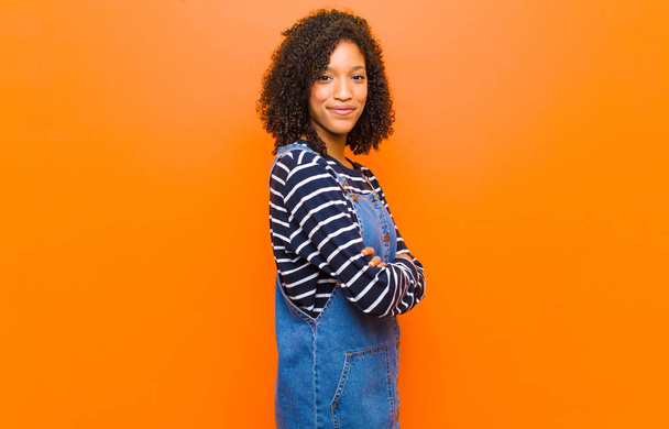 young pretty black woman smiling to camera with crossed arms and a happy, confident, satisfied expression, lateral view against orange wall - Fotoğraf, Görsel