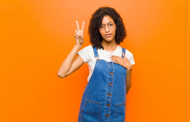 young pretty black woman looking happy, confident and trustworthy, smiling and showing victory sign, with a positive attitude against orange wall - Fotoğraf, Görsel