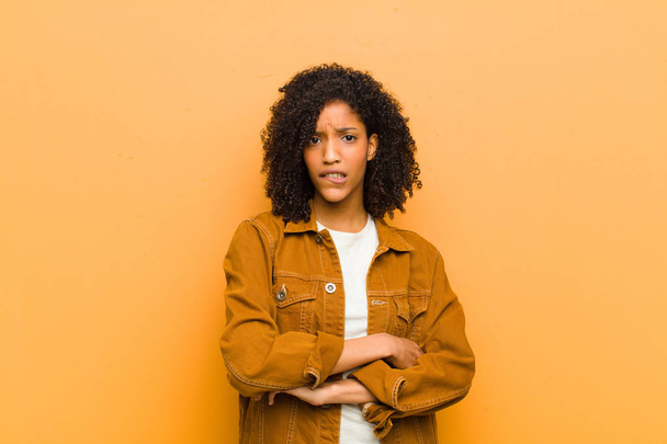 young pretty black woman doubting or thinking, biting lip and feeling insecure and nervous, looking to copy space on the side against orange wall - Zdjęcie, obraz