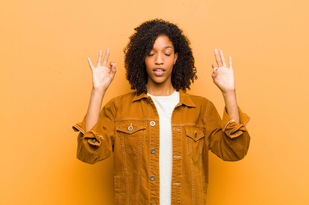 young pretty black woman looking concentrated and meditating, feeling satisfied and relaxed, thinking or making a choice against orange wall - Фото, изображение