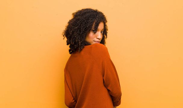 young pretty black woman smiling gleefully, feeling happy, satisfied and relaxed, with crossed arms and looking to the side against orange wall - 写真・画像