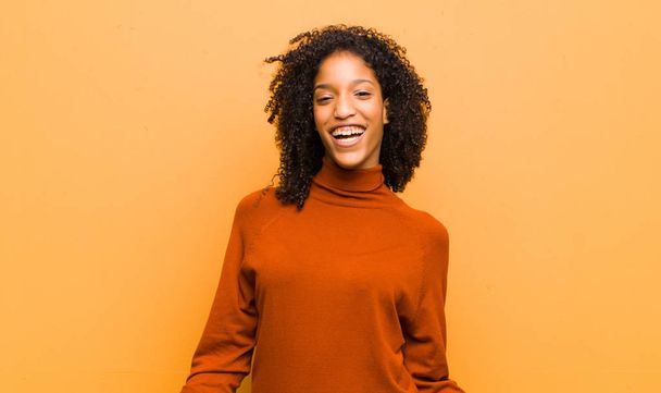 young pretty black woman looking extremely happy and surprised, celebrating success, shouting and jumping against orange wall - Photo, image