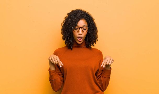 mladá hezká černá žena s otevřenými ústy ukazuje oběma rukama dolů, vypadá šokovaně, ohromeně a překvapeně proti oranžové stěně - Fotografie, Obrázek