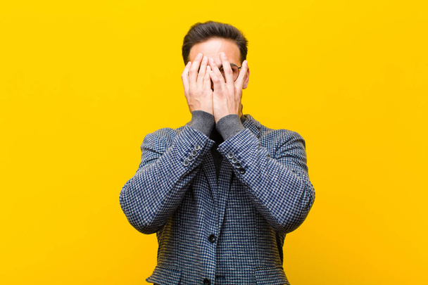 young handsome man covering face with hands, peeking between fingers with surprised expression and looking to the side against orange wall - Foto, afbeelding