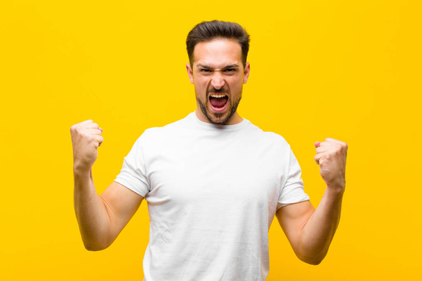 young handsome man shouting aggressively with an angry expression or with fists clenched celebrating success against orange wall - Foto, Imagen