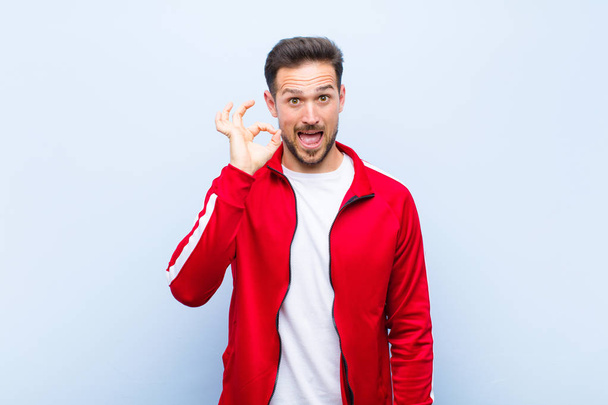 young handsome sports man or monitor feeling successful and satisfied, smiling with mouth wide open, making okay sign with hand against flat wall - Photo, image