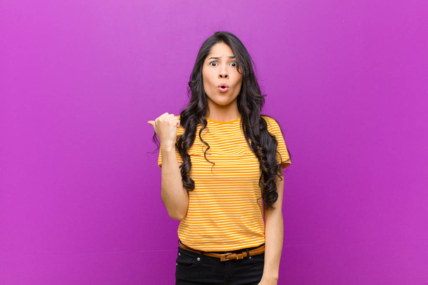 young pretty latin woman looking astonished in disbelief, pointing at object on the side and saying wow, unbelievable against purple wall - Photo, Image