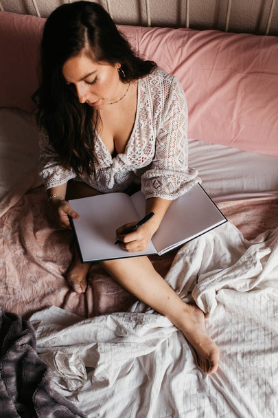 Top view of young brunette woman writing in a notebook on bed at - Foto, Imagem