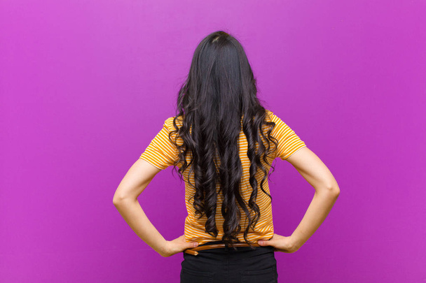 young pretty latin woman feeling confused or full or doubts and questions, wondering, with hands on hips, rear view against purple wall - Foto, Imagem