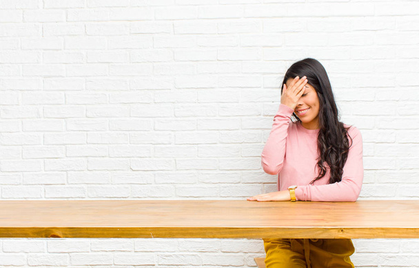 young  pretty latin woman looking stressed, ashamed or upset, with a headache, covering face with hand sitting in front of a table - Foto, Imagen