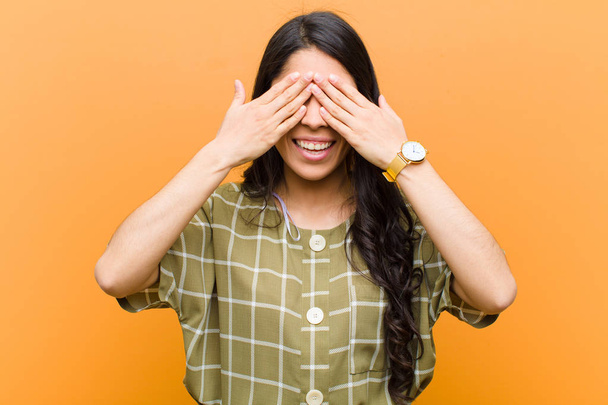 young pretty hispanic woman smiling and feeling happy, covering eyes with both hands and waiting for unbelievable surprise against brown wall - Photo, image