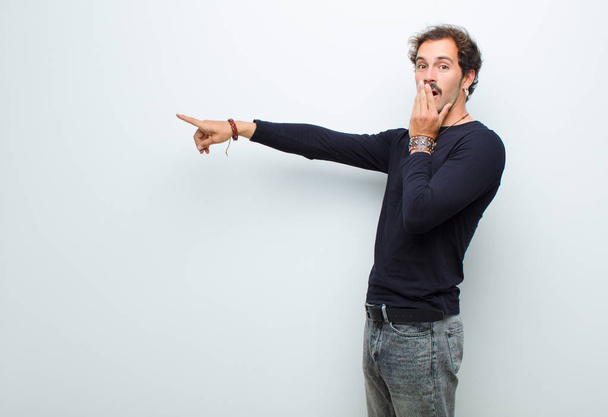 young handsome man feeling happy, shocked and surprised, covering mouth with hand and pointing to lateral copy space against white wall - Φωτογραφία, εικόνα