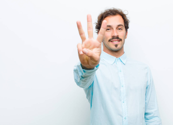 young handsome man smiling and looking friendly, showing number three or third with hand forward, counting down against white wall - Foto, Imagen