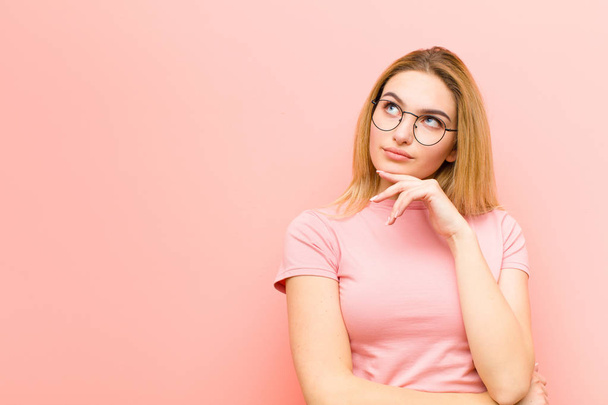 young pretty blonde woman feeling thoughtful, wondering or imagining ideas, daydreaming and looking up to copy space against pink flat wall - Foto, imagen