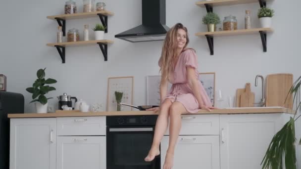 happy girl sitting on the kitchen table and carefree dangles legs - Imágenes, Vídeo