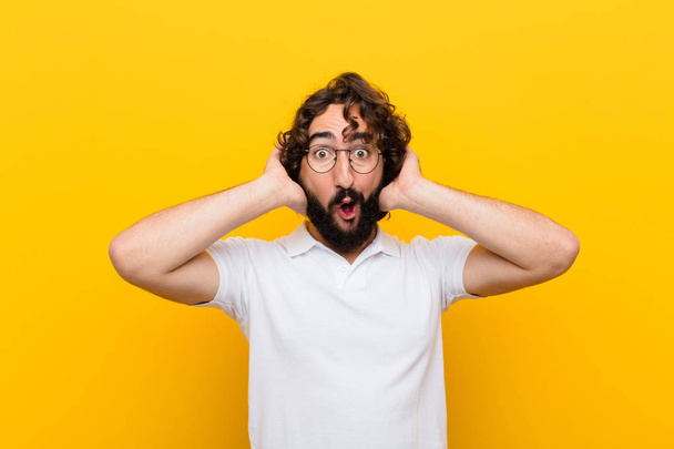 young crazy man looking excited and surprised, open-mouthed with both hands on head, feeling like a lucky winner against yellow wall - Fotoğraf, Görsel
