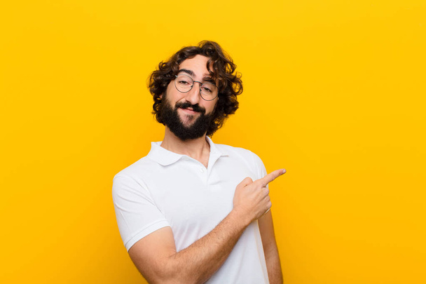 young crazy man smiling cheerfully, feeling happy and pointing to the side and upwards, showing object in copy space against yellow wall - Фото, зображення