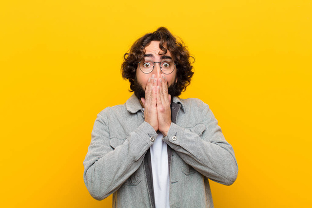 young crazy man feeling worried, upset and scared, covering mouth with hands, looking anxious and having messed up against yellow wall - Photo, image