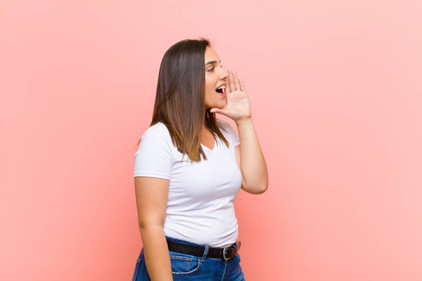 young pretty hispanic woman profile view, looking happy and excited, shouting and calling to copy space on the side against pink wall - Fotografie, Obrázek
