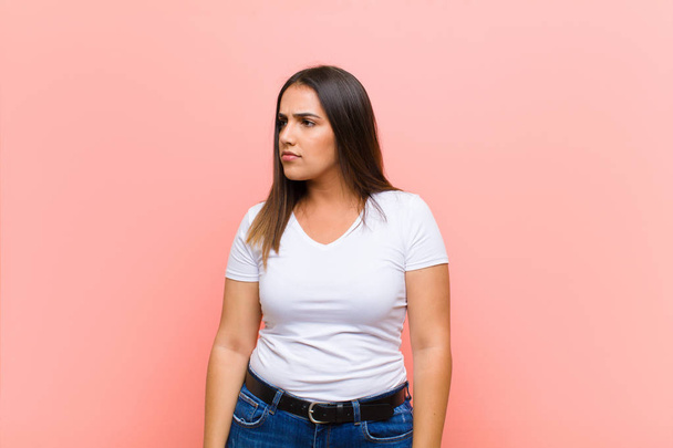 young pretty hispanic woman feeling sad, upset or angry and looking to the side with a negative attitude, frowning in disagreement against pink wall - 写真・画像